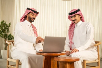 Understanding and cooperation among colleagues to achieve the company's goals, the concept of business management, completing tasks through modern devices, following up on the work strategy, two Arab Gulf men from Saudi Arabia wearing traditional attire and the ghutrah sitting in the office and working on the laptop.