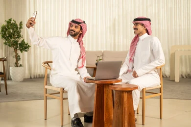Documenting happy moments, taking commemorative selfies via mobile, understanding and cooperation among colleagues to achieve the company's goals, the concept of business management, completing tasks through modern devices, following the work strategy, two Arab Gulf men from Saudi Arabia wearing traditional attire and shemagh sitting in the office and working on the laptop.
