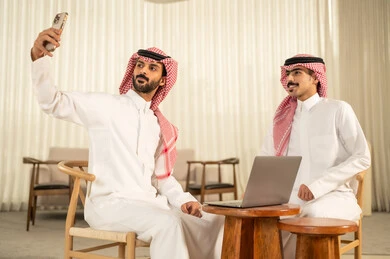 التقاط صور سيلفي التذكارية عبر الجوال ، توثيق اللحظات السعيدة ، التفاهم و التعاون بين الزملاء لتحقيق أهداف الشركة، مفهوم إدارة الأعمال، إنجاز المهام عبر الأجهزة الحديثة, متابعة إستراتيجية العمل، رجلان عربيان خليجيان سعوديان يرتديان الزي التقليدي والشماغ  يجلسان داخل المكتب و يعملان على اللابتوب