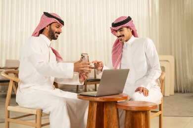 الاشارة الى شاشة الجوال ، التفاهم و التعاون بين الزملاء لتحقيق أهداف الشركة، مفهوم إدارة الأعمال، إنجاز المهام عبر الأجهزة الحديثة, متابعة إستراتيجية العمل، رجلان عربيان خليجيان سعوديان يرتديان الزي التقليدي والشماغ  يجلسان داخل المكتب و يعملان على اللابتوب