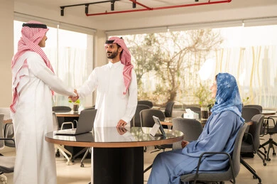 مصافحة و سلام حار بين رجلين, بيئة العمل المريحة في الشركات الراقية، مفهوم التعاون والعمل المشترك, تعابير وايماءات السعادة والسرور، رجلان عربيان خليجيان سعوديان مبتسمان يرتديان الزي التقليدي والشماغ يقفان أمام المكتب و يقومان بمصافحة بعضهم, استخدام الأجهزة المتطورة ، موظفة سعودية تجلس امام جهاز التابلت