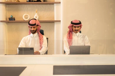 التفاهم و التعاون بين الزملاء لتحقيق أهداف الشركة، مفهوم إدارة الأعمال، إنجاز المهام عبر الأجهزة الحديثة, متابعة إستراتيجية العمل، زملاء عمل عرب خليجيين سعوديين يجلسون داخل المكتب و يعملان على اللابتوب و التابلت