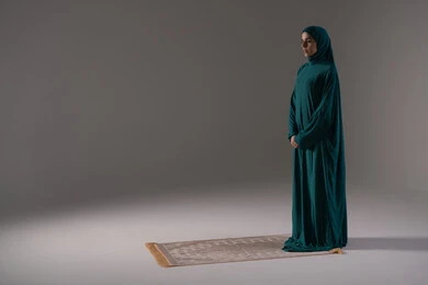 الاستعداد لاداء الصلاة, أداء الفروض على وقتها, التقرب الى الله من خلال الإجتهاد في العبادة والحفاظ على الصلوات، بورتريه  لإمرأة عربية خليجية سعودية ترتدي الحجاب والعباءة تقف على سجادة الصلاة, صورة الجسم بالطول الكامل, خلفية رمادية