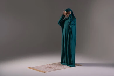 The call to prayer and preparation to start praying, worship and getting closer to God, bowing and prostration and humility, performing the obligatory prayers on time, the concept of the pillars of Islam, a portrait of a young Arab Gulf Saudi Muslim woman wearing a hijab and abaya standing on a prayer rug, gray background.