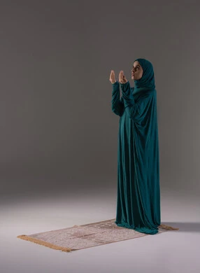 Worship and drawing closer to God, bowing and prostration and humility, supplication and pleading to the Lord of the servants, performing the obligatory acts on time, the concept of the pillars of Islam, a portrait of a young Arab Gulf Saudi Muslim woman wearing a hijab and abaya standing on a prayer rug, full-body image, gray background.