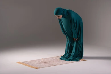 قراءة ما تيسر من القرآن الكريم، الركوع و السجود والخشوع، مفهوم أركان الإسلام ،أداء فرائض الصلاة بأوقاتها، بورتريه لإمرأة عربية خليجية سعودية مسلمة ترتدي الحجاب والعباءة تقف على السجادة، العبادة والتقرب إلى الله،خلفية رمادية