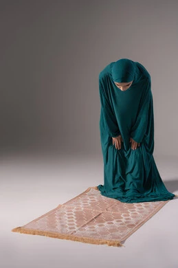 الركوع و السجود والخشوع، مفهوم أركان الإسلام ،أداء فرائض الصلاة بأوقاتها، بورتريه لإمرأة عربية خليجية سعودية مسلمة ترتدي الحجاب والعباءة تقف على السجادة، العبادة والتقرب إلى الله، قراءة ما تيسر من القرآن الكريم،خلفية رمادية