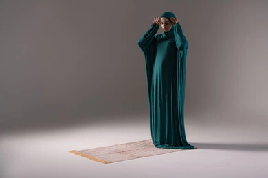 Preparation for performing prayers, fulfilling obligations on time, the concept of the pillars of Islam, a portrait of an Arab Gulf Saudi woman wearing a hijab and abaya standing on a prayer rug, worship and drawing closer to God, bowing, prostration, and humility, gray background.