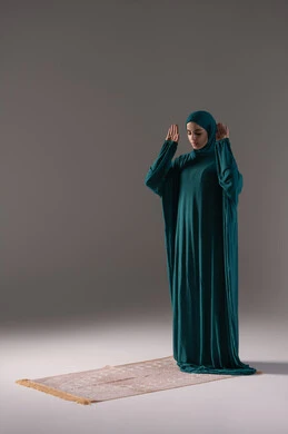 The call to prayer and preparation to start praying, performing the obligatory prayers on time, the concept of the pillars of Islam, a portrait of a Saudi Gulf Arab woman wearing a hijab and abaya standing on a prayer rug, worship and drawing closer to God, bowing, prostration, and humility, gray background.