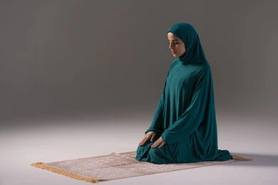 الالتزام بمواقيت الصلاة اليومية، التقرب الى الله تعالى بالإجتهاد في العبادة، بورتريه لإمرأة عربية سعودية خليجية ترتدي الحجاب والعباءة تجلس على السجادة تقوم بأداء فريضة الصلاة، مفهوم أركان الاسلام والأيمان، خلفية رمادية