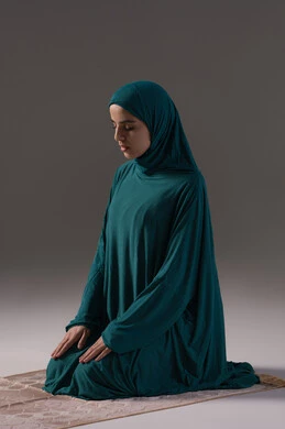 The commitment to performing the five daily prayers on time, drawing closer to God Almighty through diligence in worship, a portrait of a Saudi Arabian Gulf woman wearing a hijab and abaya sitting on a prayer rug performing the obligatory prayer, the concept of the pillars of Islam and faith, gray background.