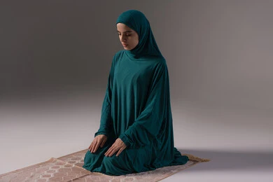Maintaining the performance of the five daily prayers on time, drawing closer to Allah Almighty through diligence in worship, a portrait of a Saudi Arabian Gulf woman wearing a hijab and abaya sitting on a prayer rug performing the obligatory prayer, the concept of the pillars of Islam and faith, gray background.