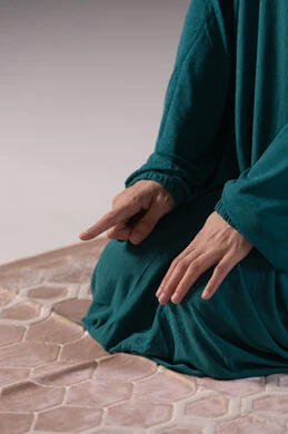 The concept of the pillars of Islam and faith, raising the index finger upwards, getting closer to God by striving in worship and maintaining prayers, a close-up portrait of the hand of a Saudi Arabian Gulf woman wearing a hijab and abaya sitting on a prayer rug performing the Tashahhud, commitment to performing the obligatory prayers on time, gray background.