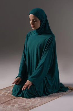 Raising the index finger upwards, getting closer to God by striving in worship and maintaining prayers, a portrait of a Saudi Arabian Gulf woman wearing a hijab and abaya sitting on a prayer rug performing the Tashahhud, the concept of the pillars of Islam and faith, commitment to performing the obligatory prayers on time, gray background.