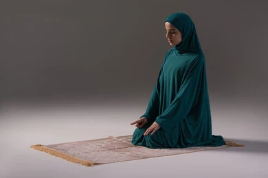 Drawing closer to God through diligence in worship and maintaining prayers, a portrait of a Saudi Arabian Gulf woman wearing a hijab and abaya sitting on a prayer rug performing the Tashahhud, raising her index finger upwards, the concept of the pillars of Islam and faith, commitment to performing obligatory prayers on time, gray background.
