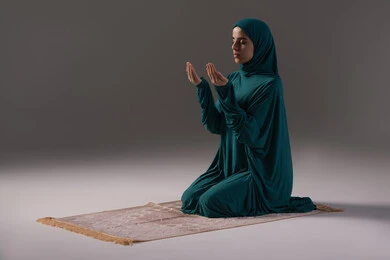 التوسل إلى الله بالرجاء والخضوع، طلب العون والرغبة في تحقيق الأمنيات, بورترية لامراة عربيه خليجه عربيه سعودية ترتدي العباءة و الحجاب تجلس على سجادة صلاه تنظر إلى الأعلى ترفع يديها بخشوع واستعطاف, خلفية رمادية