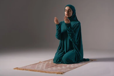 Turning to God in prayer and humility, seeking help and desiring to fulfill wishes, a portrait of an Arab Gulf woman, a Saudi Arabian, wearing an abaya and hijab, sitting on a prayer rug, looking up, raising her hands in reverence and supplication, with a gray background.