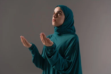 Requesting help and the desire to fulfill wishes, supplication to God Almighty, prayer, intercession, and seeking mercy, asking with hope and humility from the Lord of the worlds, a portrait of an Arab Gulf woman, a Saudi Arabian woman wearing an abaya and hijab, looking up, raising her hands in reverence and pleading, with a gray background.