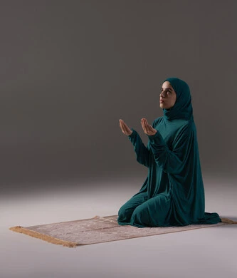 التضرع إلى الله تعالى, طلب العون والرغبة في تحقيق الأمنيات, الدعاء والتوسل والاسترحام, الطلب بترجي وتذلل من رب العباد, بورترية لامراة عربيه خليجيه عربيه سعودية ترتدي العباءة و الحجاب تجلس على سجادة الصلاة تنظر إلى الأعلى ترفع يديها بخشوع واستعطاف, خلفية رمادية