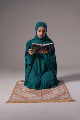 المناسبات الدينية والأعياد الإسلامية، التفكر والخشوع في التقرب إلى الله، بورترية لامراة عربيه خليجه عربيه سعودية ترتدي العباءة و الحجاب تجلس علي سجادة الصلاة و تقرأ القرآن الكريم، خلفيه رمادية