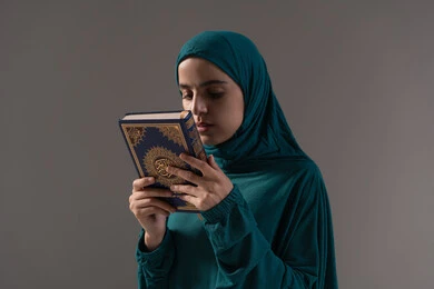 الاجتهاد في العباده والطاعة، تعظيم المصحف الشريف، حب كتاب الله و تقبيله, بورترية لامراة عربيه خليجه عربيه سعودية ترتدي العباءة و الحجاب تمسك القرآن الكريم بيديها، خلفية رمادية