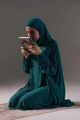 حب كتاب الله و تقبيله، تعظيم المصحف الشريف, بورترية لامراة عربيه خليجه عربيه سعودية ترتدي العباءة و الحجاب  تجلس على سجادة الصلاة تمسك القرآن الكريم بيديها، الاجتهاد في العباده والطاعة، خلفية رمادية
