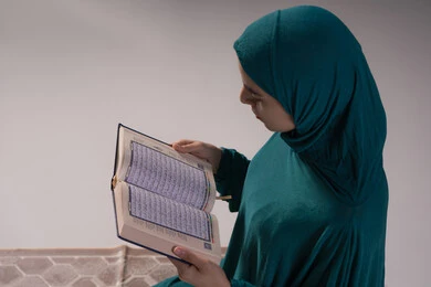 الخشوع والتمعن في أداء العبادات، التقرب الى الله تعالى، بورترية مقرب من الجانب لامراة عربيه خليجه عربيه سعودية ترتدي العباءة و الحجاب تقرأ القرآن الكريم، المناسبات الدينية والأعياد الإسلامية، خلفيه رمادية