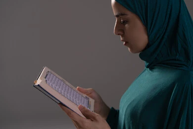 التدبر في معاني العبادة والخشوع لها، التقرب الى الله تعالى، بورترية مقرب من الجانب لامراة عربيه خليجه عربيه سعودية ترتدي العباءة و الحجاب تقرأ القرآن الكريم، المناسبات الدينية والأعياد الإسلامية، خلفيه رمادية