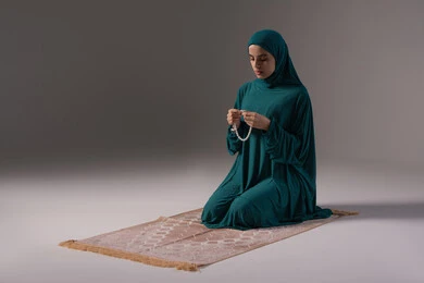 ترديد الأدعية وذكر الله المستمر بالاستغفار، الإجتهاد في العباده والتقرب الى الله في شهر رمضان, مفهوم التسبيح والتهليل، بورترية لامراة عربيه خليجه عربيه سعودية ترتدي العباءة و الحجاب تجلس على سجادة الصلاة تمسك بيدها السبحه, خلفيه رمادية