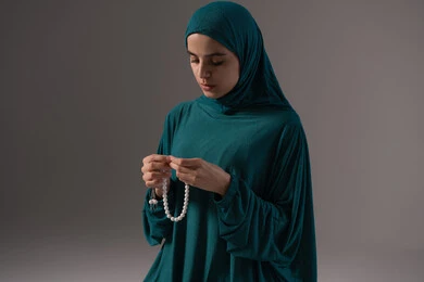 الإجتهاد في العباده والتقرب الى الله في شهر رمضان, مفهوم التسبيح والتهليل، بورترية لامراة عربيه خليجه عربيه سعودية ترتدي العباءة و الحجاب  تمسك بيدها السبحه, الإكثار من ذكر الله تعالى من خلال الإستغفار والدعاء, خلفيه رمادية