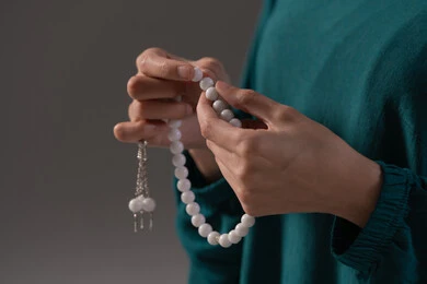 The concept of glorification and declaration of faith, striving in worship and drawing closer to God in the month of Ramadan, a close-up portrait of the hand of an Arab Gulf woman, a Saudi Arabian woman wearing an abaya holding a prayer bead, increasing the remembrance of God Almighty through seeking forgiveness and supplication, gray background.