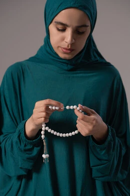 The abundance of mentioning Allah Almighty through seeking forgiveness and supplication, striving in worship and getting closer to Allah during the month of Ramadan, the concept of glorification and declaration of faith, a portrait of an Arab Gulf woman, a Saudi Arabian woman wearing an abaya and hijab holding a prayer bead in her hand, with a gray background.