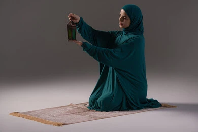 الاستعداد للعبادات والأجواء الرمضانية، بورترية لامراة عربيه خليجه عربيه سعودية ترتدي العباءة و الحجاب تجلس على سجادة الصلاة تحمل بيدها فانوس رمضان الملون, ديكورات شهر رمضان المبارك، خلفية رمادية