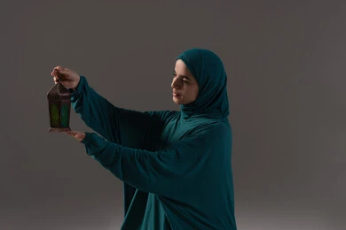 Preparing oneself and the places for the holy month, a portrait of an Arab Gulf woman, a Saudi Arabian, wearing an abaya and hijab, sitting on a prayer rug holding a colorful Ramadan lantern in her hand, decorations for the blessed month of Ramadan, gray background.