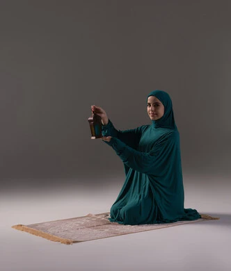 تعابير السعادة و السرور، الاحتفال بالمناسبات الدينية الإسلامية، التحضير لاستقبال رمضان المبارك, البهجة بقدوم شهر الخير, بورتريه لامرأة عربية خليجية سعودية محجبة ترتدي العباءة تجلس على سجادة الصلاة تمسك بيدها فانوس معدني مزخرف، خلفية رمادية