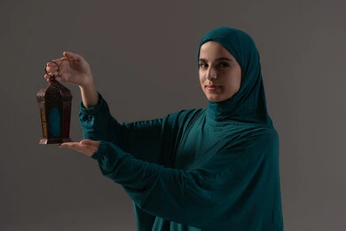 البهجة بقدوم شهر الخير، التحضير لاستقبال رمضان المبارك, بورتريه  لامرأة عربية خليجية سعودية محجبة ترتدي العباءة تمسك بيدها فانوس معدني مزخرف, الاحتفال  بالمناسبات الدينية الإسلامية, تعابير السعادة و السرور، خلفية رمادية