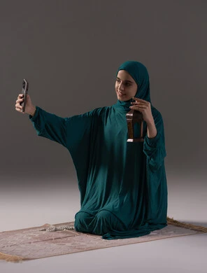 The atmosphere of tranquility and spirituality, religious occasions and Islamic holidays, gestures of happiness and joy, a portrait of a Saudi Arabian Gulf woman wearing a hijab and abaya holding a mobile phone taking a picture of a metal lantern, gray background.