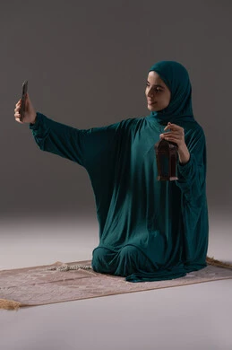 الخشوع والطاعة والتقرب لله تعالى، الاستمتاع بالروحانية الرمضانية وأجواء العبادة، ايماءات السعادة و السرور، بورتريه لإمرأة عربية سعودية خليجية ترتدي الحجاب والعباءة تمسك بيدها الهاتف المحمول تقوم بالتقاط صورة لفانوس معدني، خلفية رمادية