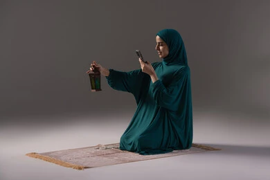 Enjoying the Ramadan spirituality and the atmosphere of worship, gestures of happiness and joy, worship and drawing closer to God, a portrait of a Saudi Arabian Gulf woman wearing a hijab and abaya holding a mobile phone taking a picture of a metal lantern, gray background.