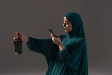 قضاء أوقات روحانية في الشهر الفضيل، ايماءات السعادة و السرور، العبادة والتقرب إلى الله، بورتريه لإمرأة عربية سعودية خليجية ترتدي الحجاب والعباءة تمسك بيدها الهاتف المحمول تقوم بالتقاط صورة لفانوس معدني، خلفية رمادية قضاء أوقات روحانية في الشهر الفضيل، ايماءات السعادة و السرور، العبادة والتقرب إلى الله، بورتريه لإمرأة عربية سعودية خليجية ترتدي الحجاب والعباءة تمسك بيدها الهاتف المحمول تقوم بالتقاط صورة لفانوس معدني، خلفية رمادية