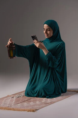 ايماءات السعادة و السرور، العبادة والتقرب إلى الله، الاستمتاع بالأجواء الروحانية في شهر رمضان المبارك، بورتريه لإمرأة عربية سعودية خليجية ترتدي الحجاب والعباءة تمسك بيدها الهاتف المحمول تقوم بالتقاط صورة لفانوس معدني، خلفية رمادية