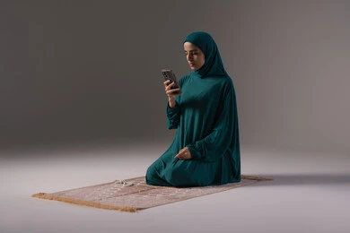 ترديد الأذكار ورفع الأدعية إلى الله، مفهوم التطبيقات والبرامج الدينية، بورتريه لإمرأة عربية سعودية اماراتية ترتدي الحجاب والعباءة تجلس على سجادة الصلاة تمسك بيدها الهاتف المحمول تقوم بتلاوة القرآن من المصحف الالكتروني عبر تطبيقات الجوال، خلفية رمادية