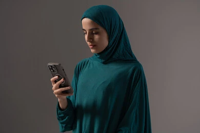 Reading the remembrances and supplications, the concept of religious applications and programs, a portrait of an Arab Saudi Emirati woman wearing a hijab and abaya holding a mobile phone reciting the Quran from the electronic mushaf through mobile applications, gray background.