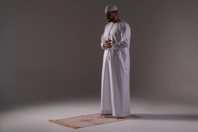 الإخلاص في الطاعة والتعبد لله، أداء الصلوات على وقتها، الشعور بالخشوع والطمأنينة والسكون , بورتريه لرجل عربي خليجي إماراتي يرتدي الكندورة و القبعة البيضاء يقف باستقامة على السجادة يؤدي فريضة الصلاة، الركن الثاني من أركان الإسلام، خلفية رمادية
