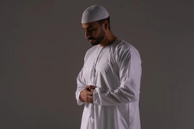 السعي للتقرب إلى الله بالعبادات، أداء الصلوات على وقتها، الشعور بالخشوع والطمأنينة والسكون , بورتريه لرجل عربي خليجي إماراتي يرتدي الكندورة و القبعة البيضاء يقف باستقامة على السجادة يؤدي فريضة الصلاة، الركن الثاني من أركان الإسلام، خلفية رمادية