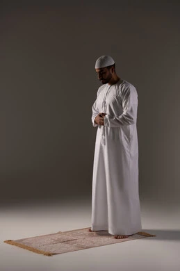Obedience and humility to Allah Almighty, performing prayers on time, feeling of humility, tranquility, and calmness. A portrait of an Emirati Gulf Arab man wearing a kandura and a white cap standing upright on the prayer rug performing the obligatory prayer, the second pillar of Islam, with a gray background.