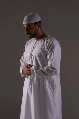 Performing prayers on time, feeling humility, tranquility, and calmness, worship and drawing closer to God, a portrait of an Emirati Gulf Arab man wearing a kandura and a white cap standing upright on the prayer rug performing the obligatory prayer, the second pillar of Islam, with a gray background.