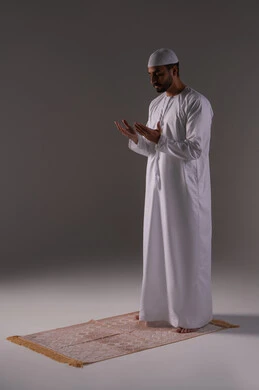 The request is made with pleading and humility to the Lord of the worlds, standing with gestures of prayer, supplication, and seeking mercy, asking for help and desiring to fulfill wishes, supplicating to God Almighty. A portrait of an Arab Gulf Emirati man wearing a kandura and a white cap, standing on a prayer rug, raising his hands with reverence and pleading, with a white background.