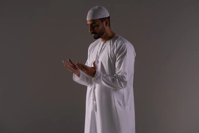 Standing with gestures of prayer, supplication, and seeking mercy, asking for help and desiring to fulfill wishes, pleading to God Almighty. A portrait of an Emirati Arab man wearing a kandura and a white cap, raising his hands in humility and pleading, requesting with entreaty and submission from the Lord of the worlds, with a white background.