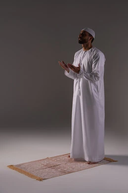 التضرع إلى الله تعالى, بورتريه لرجل عربي خليجي إماراتي يرتدي الكندورة و القبعة البيضاء يقف على سجادة الصلاة يرفع يديه بخشوع واستعطاف, طلب العون والرغبة في تحقيق الأمنيات, الوقوف بإيماءات الدعاء والتوسل والاسترحام, الطلب بترجي وتذلل من رب العباد, خلفية بيضاء